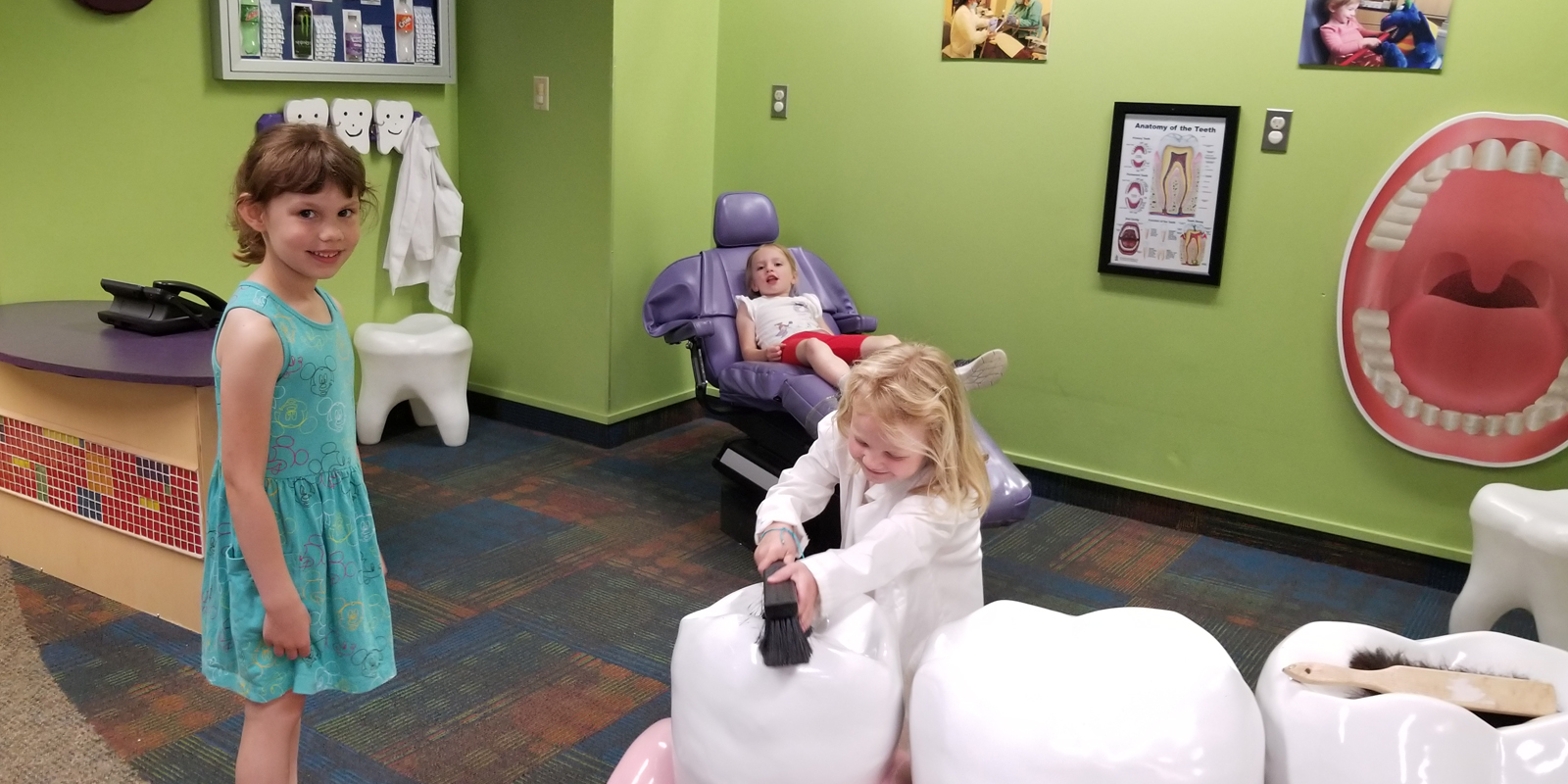 Three young girls playing with giant plastic teeth, toothbrush and dental chair.