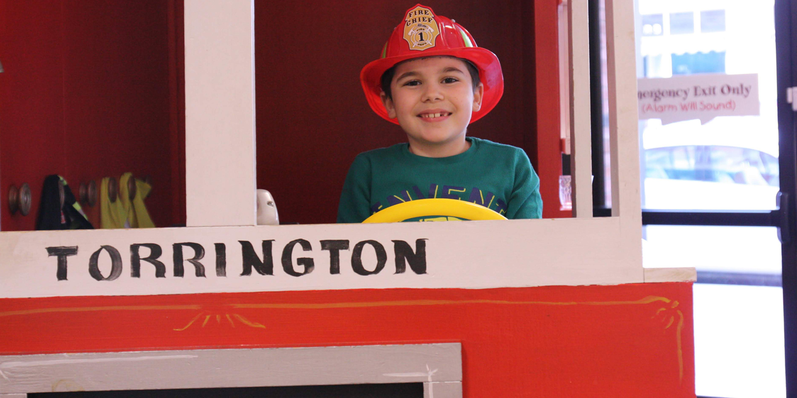 Young boy wearing a plastic fire hat and driving a play wooden fire truck