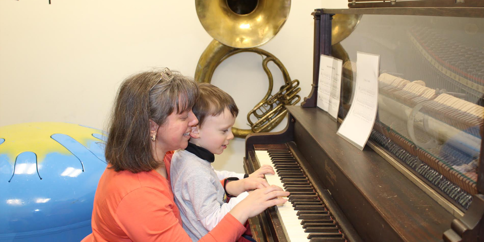 Woman with light brown hair wearing orange shirt is helping young boy play the piano