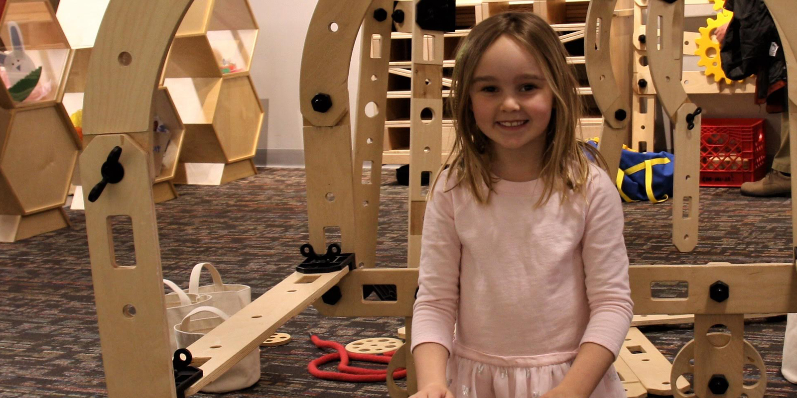 Young girl with pink dress is smiling in front of large wooden erector set