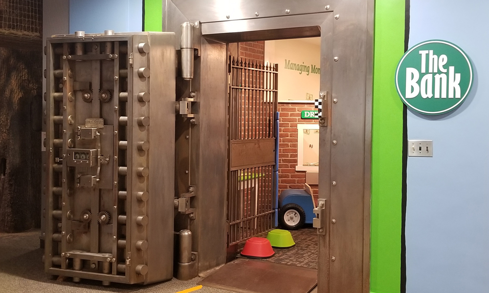 Exhibit of bank vault with green trim and a heavy looking vault door open.