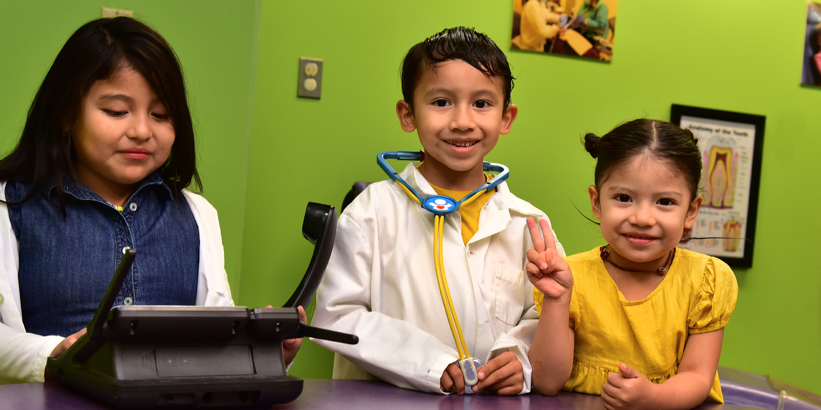 Three young children smiling in a room with bright green walls. Young boy wearing a play doctor outfit.