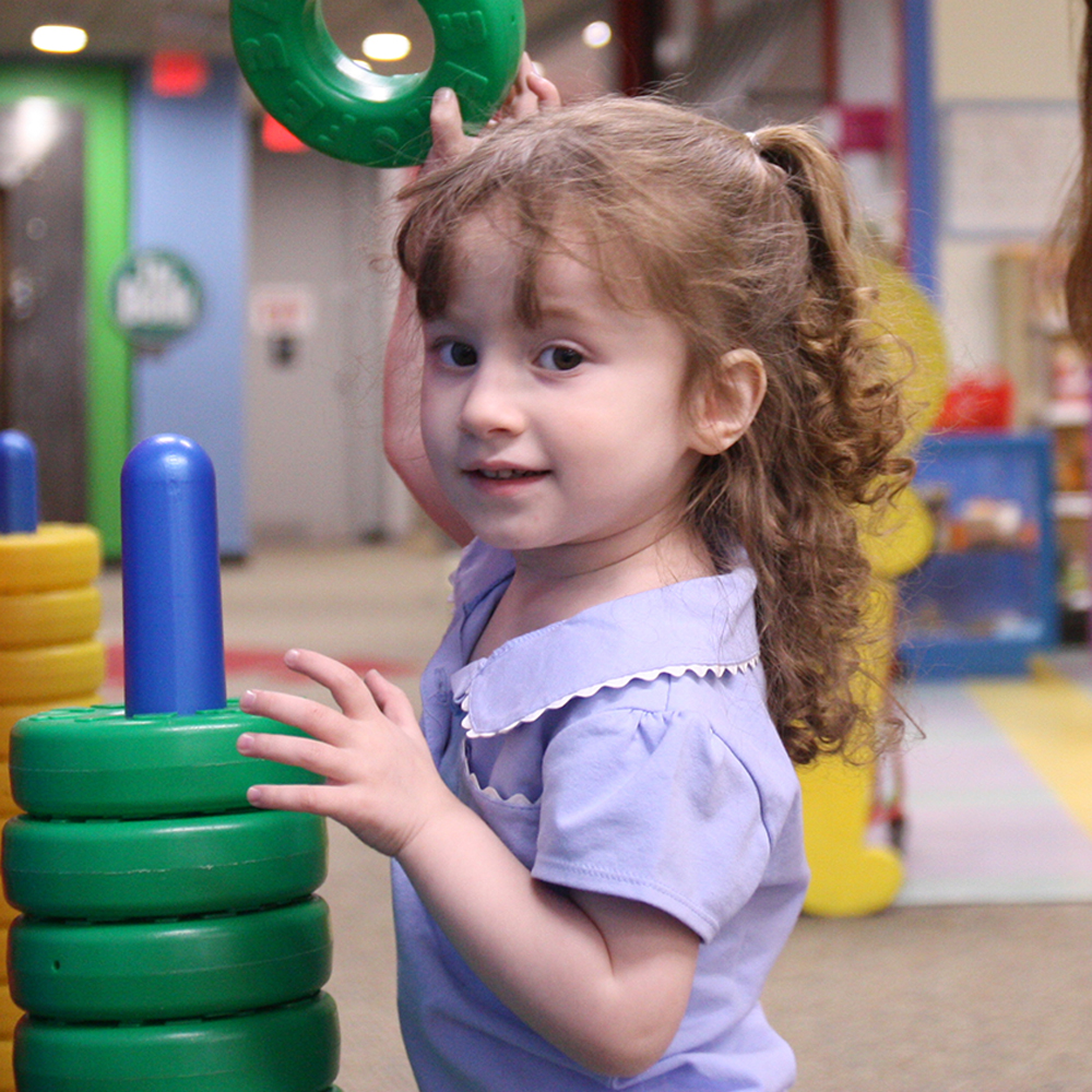 Young girl with light brown curly hair in ponytail putting green plastic rings on a blue pole
