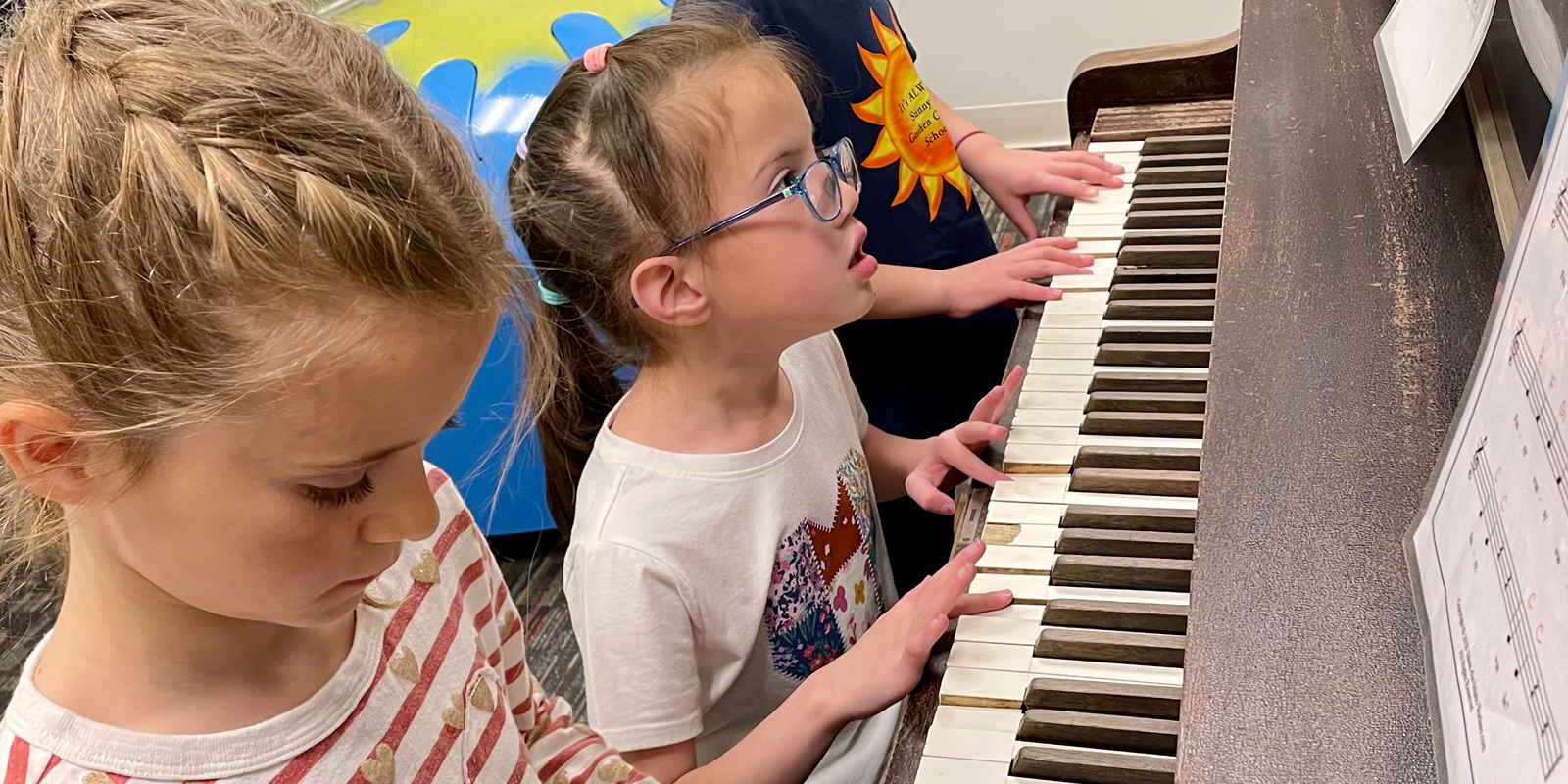 Young girl with blue-rimmed glasses and ponytail in between two other children playing piano