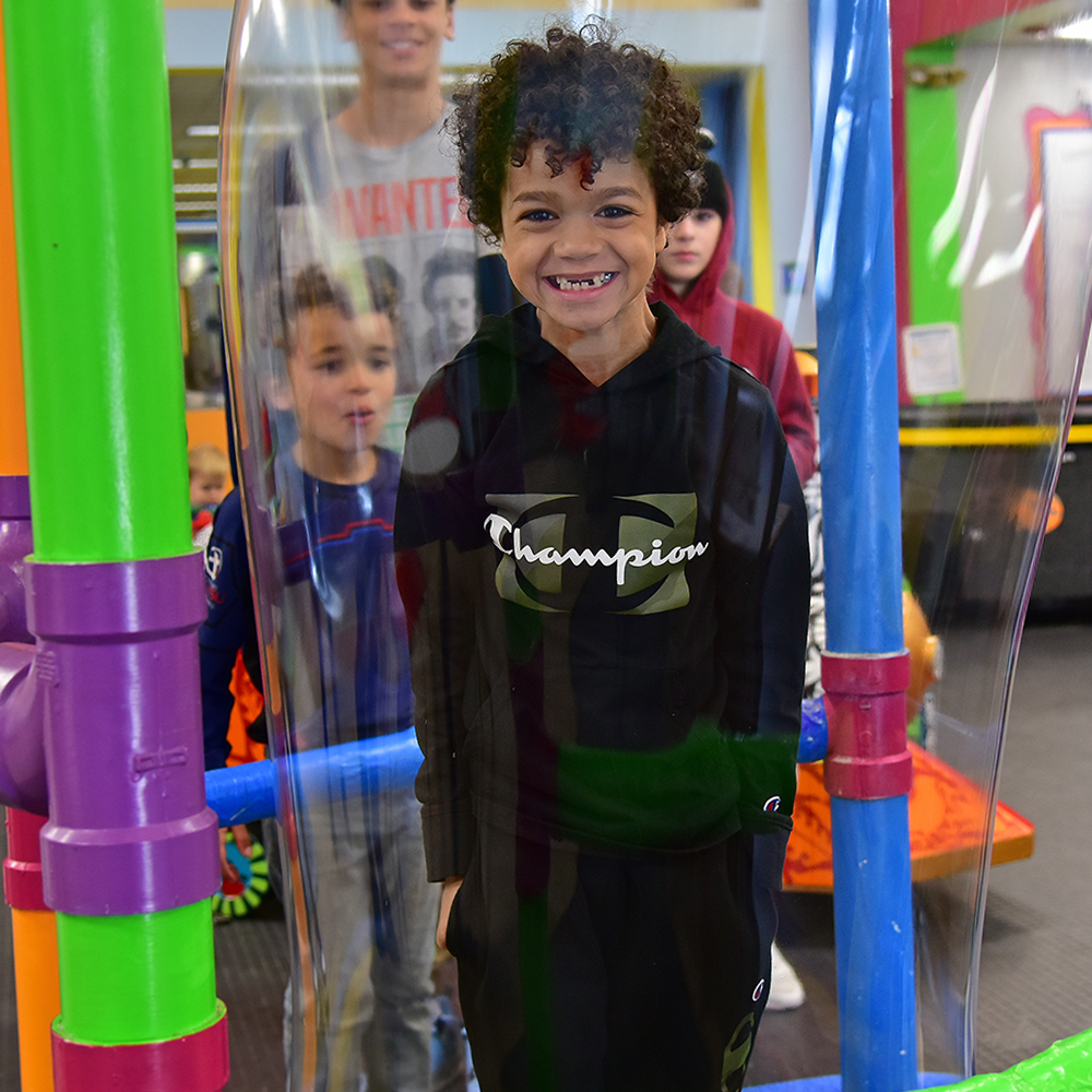Young boy with dark curly hair smiling inside of a large bubble with other kids watching
