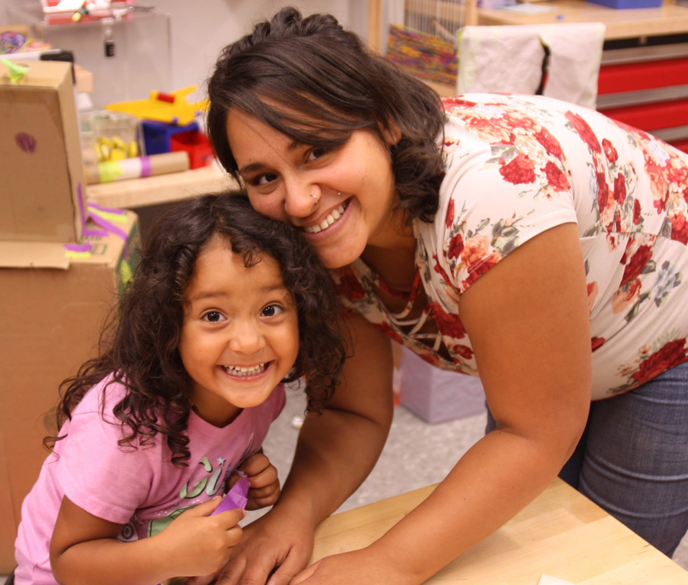 Young woman in white flowered shirt and jeans building paper crafts with young girl with dark hair and pink t-shirt.
