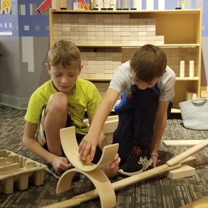 A photo of two boys playing with arch-shaped blocks in KidsPlay's Block City exhibit. The boy on the left wears a yellow shirt and the boy on the right wears a white shirt. Neither boy is looking directly at the camera; instead, both are smiling slightly and seem deeply absorbed in their play.