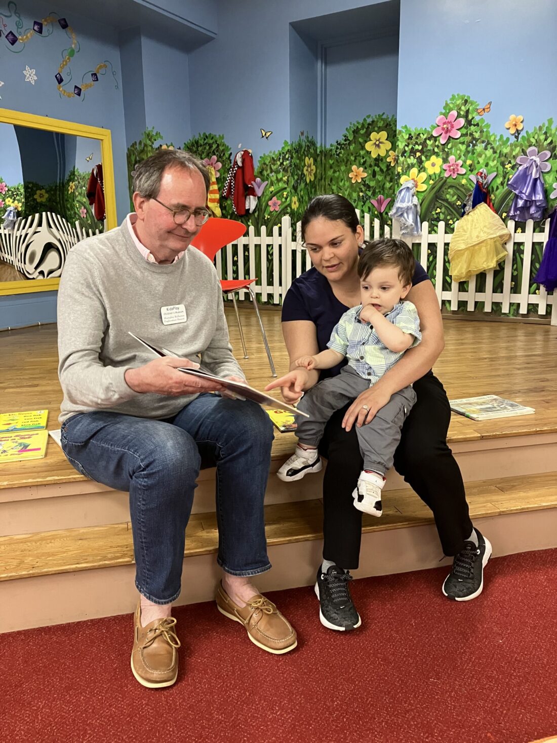 A photo of three people sitting on the stairs of the KidsPlay stage. A volunteer (an older man wearing blue jeans, glasses, and a gray sweater) is reading a picture book. A younger woman in black clothes holds a young child, who is pointing at the picture book.