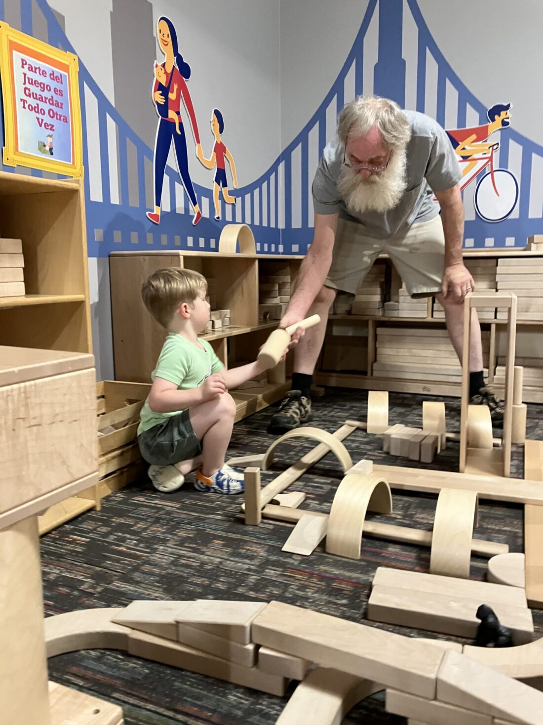 A photo of an older man with a white beard and a young boy in a green shirt playing together in KidsPlay's Block City area. The older man is stooping down and handing the young boy a cylindrical block.