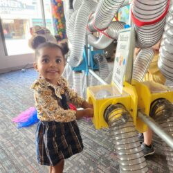 A photo of a young girl playing with the Air Tubes exhibit at KidsPlay Children's Museum. She is smiling and looking up at the camera.