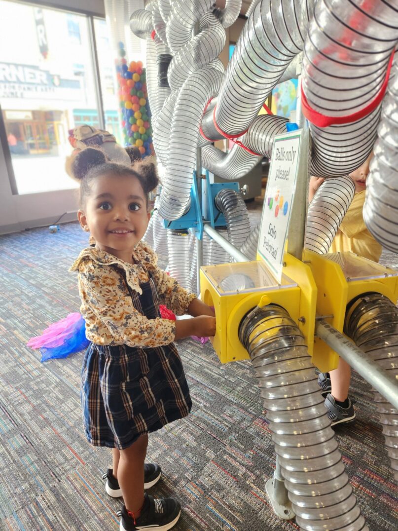 A photo of a young girl playing with the Air Tubes exhibit at KidsPlay Children's Museum. She is smiling and looking up at the camera.