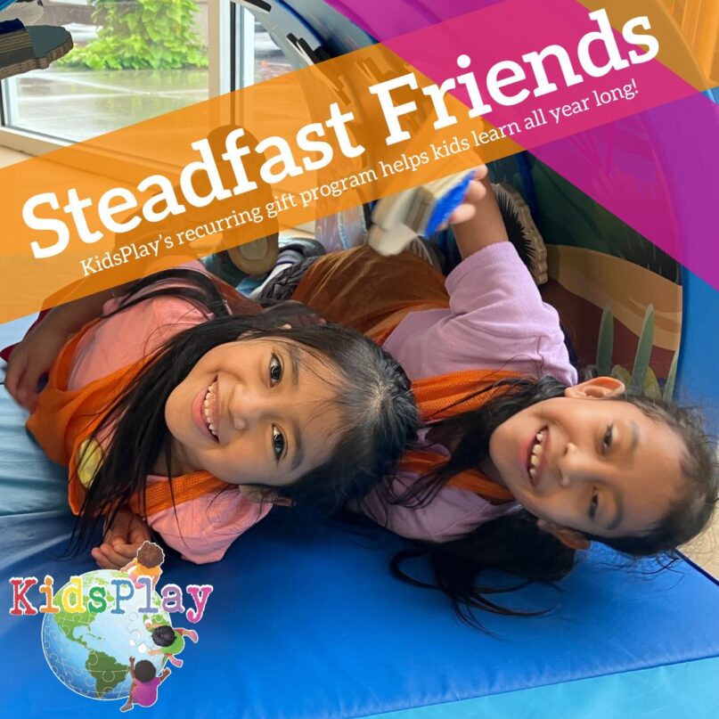 A square graphic with white text on a transparent orange background reading "Steadfast Friends" with a subheading reading "KidsPlay's recurring gift program helps kids learn all year long!" The background is a photo of two girls playing at KidsPlay. Both are laying on their side on the mat in the Freeman Foundation Asian Culture Exhibit "Play" and smiling at the camera. The KidsPlay logo is in the lower left hand corner of the graphic. The text and the orange stripe are on an angle above the girls' heads, and the orange stripe is intersected by a transparent hot pink stripe running in another direction.