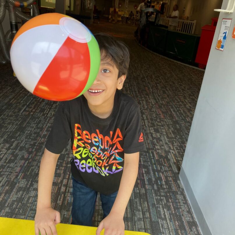 A photo of a young boy playing with the Bernoulli Box exhibit at KidsPlay. One of the floating balls partially obscures his face. He is smiling and focused on the ball.