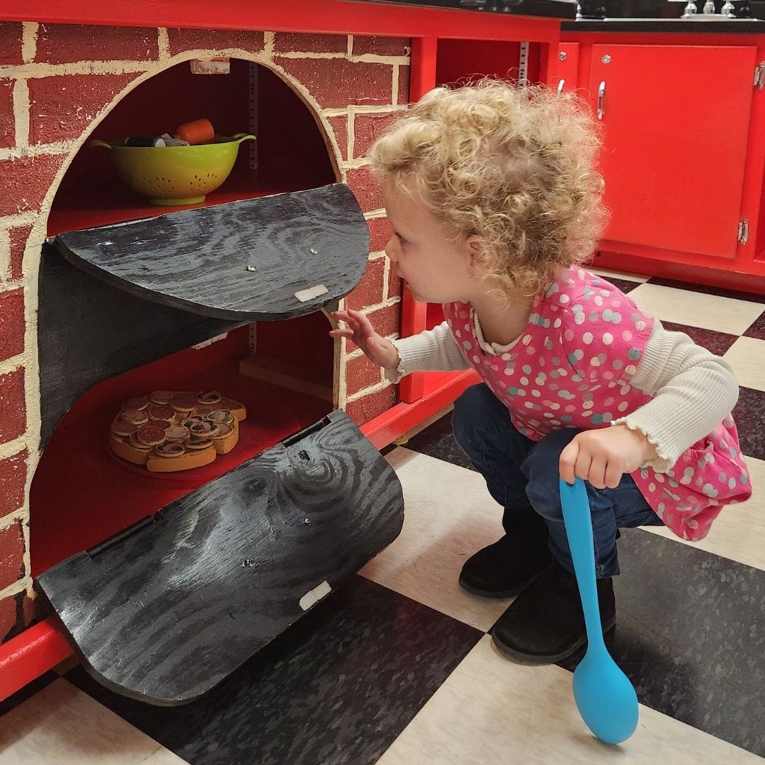 A photo of a young girl in a pink dress crouching to look into the oven in the KidsPlay Diner exhibit at KidsPlay Children's Museum in Torrington, CT.