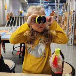 A picture of a young girl in a yellow shirt playing in the Maker Space at KidsPlay Children's Museum in Torrington, CT. She holds a pair of toy binoculars that she has made out of tissue paper and cardboard.