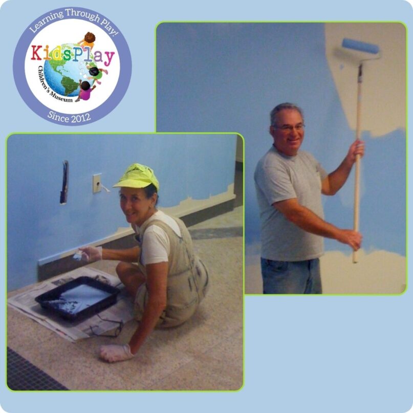 A square graphic with a blue background and a white border. There are two square-framed overlapping photos of Stu and Jane Grodin painting blue walls in the Main Gallery of KidsPlay Children's Museum in Torrington, CT.